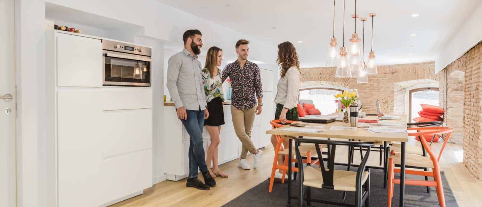 Quatre jeunes adultes dans une cuisine ouverte sur une salle à manger, mur de brique et suspensions au-dessus de la table.
