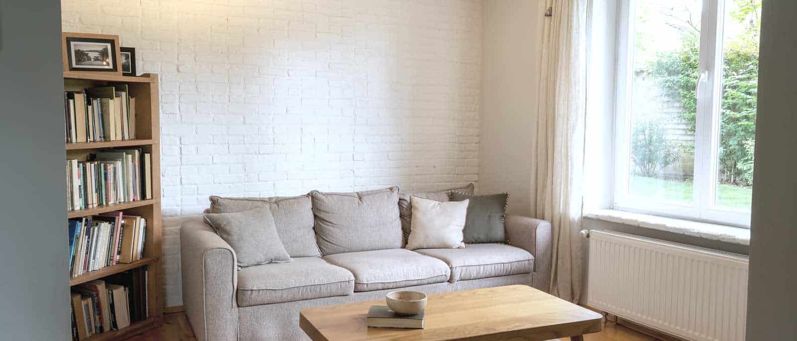 Le salon d’une maison en location, mur de brique peint en blanc, canapé clair, bibliothèque et table basse devant une fenêtre ouverte sur un jardin.